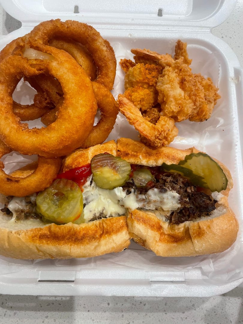 Cheesesteak sandwich with pickles, ketchup, onion rings, and fried shrimp in a white takeout container.