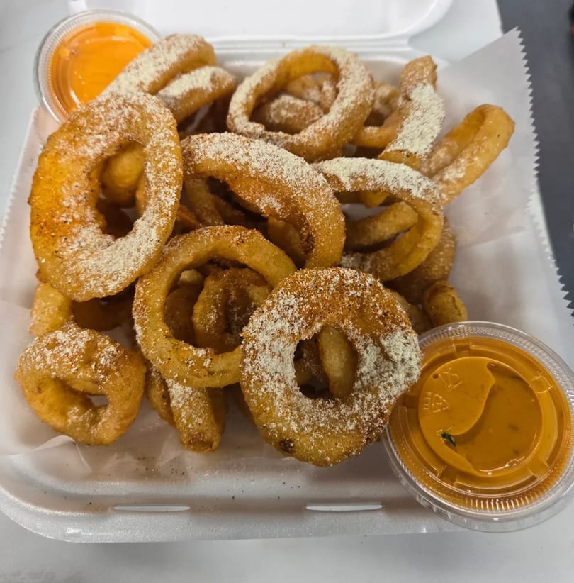 Curly fries and onion rings, dusted with seasoning, served in a white container with two dipping sauces.